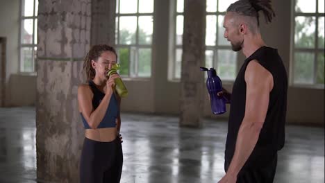 Stylish-sporty-man-and-woman-standing-in-studio-sport-club-with-windows-background-and-talking.-They-are-drinking-water-after-workout-with-color-bottles.-Both-with-stylish-dreadlocks
