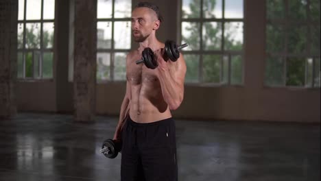 Shirtless-muscular-man-doing-workout-with-dumbbells-and-sweat-on-his-body.-Man-shirtless-with-sweaty-torso-exercising-in-empty-loft-studio.-Slow-motion