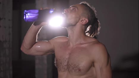 Bodybuilding-man-with-dreadlocks-portrait-standing-in-front-of-the-camera-shirtless-drinking-sensually-water-from-blue-bottle-in-lights-spotlight-of-background.-Slow-motion