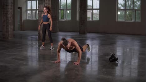 Beautiful-young-sports-couple-is-working-out-together-in-studio-gymwith-panoramic-window.-Girl-with-dreadlocks-does-squats,-man-bending-push-ups-next-to-her