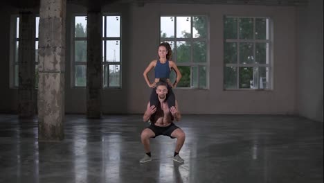 Athletic-shirtless-man-crouches-with-a-girl-on-his-shoulders-in-empty-loft-studio.-Does-squats-with-his-partner-on-shoulders.-Slow-motion.-Front-view