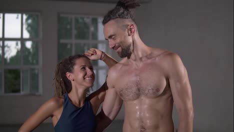 Happy-couple-with-dreadlocks-after-successful-training-in-sports-club-together.-Sport-woman-looking-on-her-shirtless-handsome-partner-or-trainer,-smiling-to-each-other-embracing