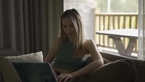 Blonde-woman-sitting-on-sofa-in-country-house,-browsing-social-media-on-laptop