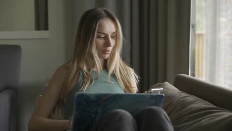 Blonde-woman-sitting-on-sofa-with-laptop-on-knees-using-her-cell-phone
