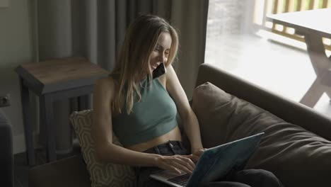Blonde-woman-sitting-on-sofa-with-laptop-and-talking-by-smartphone