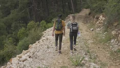 A-happy-couple-backpackers-walking-by-forest's-rocky-path,-rear-view