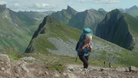 Bonita-Excursionista-Rubia-Subiendo-Una-Colina-Empinada-Frente-A-Un-Impresionante-Telón-De-Fondo-De-Montañas-Y-Fiordos,-En-Un-Soleado-Día-De-Verano-En-El-Norte-De-Noruega,-Senja