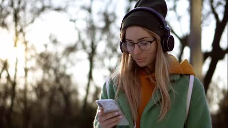 Stylish-girl-walk-in-winter-park,-listening-to-music-on-the-phone-using-headphones