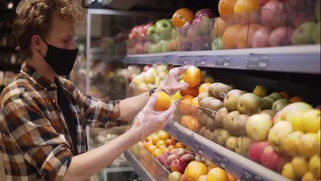 Young-man-in-plaid-shirt,-wearing-tissue-black-mask-and-plastic-transparent-gloves,-shopping-for-fruits-in-the-supermarket-during-the-quarantine-pandemic-covid-19-coronavirus.-Side-view
