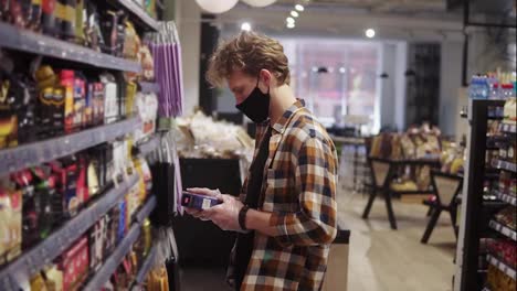 Customer-in-the-supermarket-during-pandemic-time.-Man-reading-the-label-on-the-pack-of-the-coffee.-Shelves-on-the-background.-Wearing-black-protective-mask