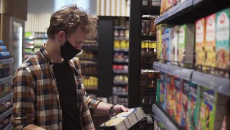 Male-customer-in-the-supermarket-during-pandemic-time.-Man-reading-the-label-on-the-pack-of-pasta.-Shelves-and-department-row-on-the-background.-Wearing-black-protective-mask-and-gloves