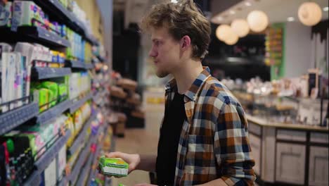 Caucasian-young-man-holding-feminine-pads-in-the-mall-and-reading-the-label,-grimacing.-Concept-of-shopping-and-choices.-Side-view.-Slow-motion