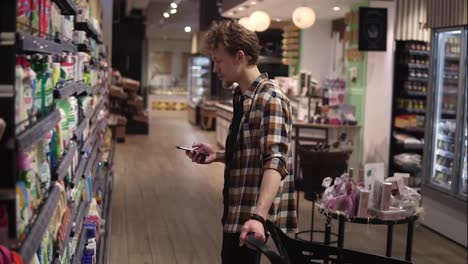 Young-man-using-smartphone-in-supermarket.-Caucasian-man-in-plaid-shirt-holding-mobile-phone-and-choosing-goods-in-grocery-store,-handheld-shot.-Shopping-and-technology-concept