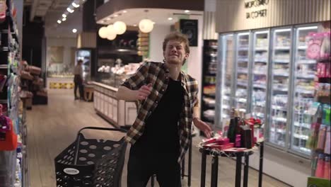 Excited-young-man-in-plaid-shirt-in-supermarket-dancing,-funky-moves-close-to-shopping-trolley.-Positive-dances-in-an-empty-food-store-row.-Positive-emotions
