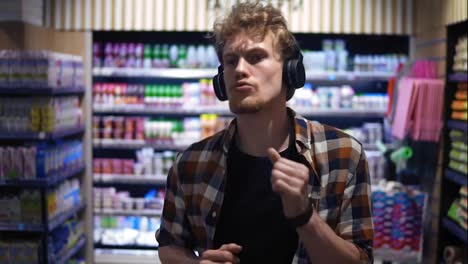 At-the-Supermarket:-Stylish-caucasian-guy-with-headphones-walks-through-goods-section-of-the-store-and-listening-to-the-music,-wearing-plaid-shirt.-Front-view.-Slow-Motion