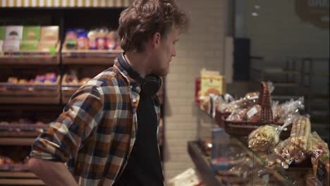 Young-guy-with-headphones-on-neck-choosing-tasty-cakes,-choosing-pastries-on-glass-showcase-in-modern-supermarket-bakery.-Side-view