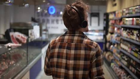 Stylish-caucasian-guy-with-headphones-walks-through-goods-section-of-the-store,-wearing-plaid-shirt---listening-to-the-good-music..-Following-back-view-shot.-Slow-Motion