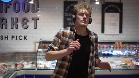 Cheerful-caucasian-guy-with-headphones-walks-through-goods-section-of-the-store-and-listening-to-the-music,-and-funny-dance-wearing-plaid-shirt.-Front-view.-Slow-Motion