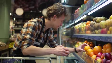 Im-Supermarkt:-Hübscher-Lagerverkäufer-Mit-Schwarzer-Schürze,-Der-Bio-Obst-Und--Gemüse-Arrangiert.-Hinzufügen-Von-Frischen-Orangen-Zum-Regal.-Zeitlupe