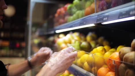 Close-up-of-shop-assistants-in-transparent-gloves-filling-up-lemons-storage-stands-with-assorted-organic-groceries-in-supermarket.-Supermarket-clerks-placing-fruits-on-display-store-shelves