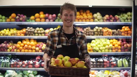 Portrait-shot-of-the-young-handsome-Caucasian-shop-worker-in-the-apron-standing-in-front-the-camera-and-smiling-joyfully-while-holding-box-of-apples-at-the-grossery-supermarket.-Fruits-and-vegetables-panel-background