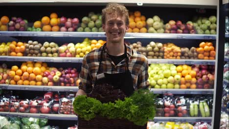 Portrait-shot-of-the-young-handsome-Caucasian-shop-worker-in-the-apron-standing-in-front-the-camera-and-smiling-joyfully-while-holding-box-coloured-greens-at-the-grossery-supermarket.-Fruits-and-vegetables-panel-background