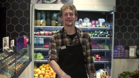Portrait-shot-of-the-young-handsome-Caucasian-shop-worker-in-the-apron-in-front-the-camera-and-smiling-joyfully-while-pushing-cart-at-the-grossery-supermarket