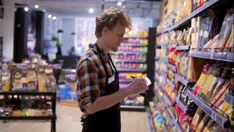Serious-store-clerk-working-at-the-supermarket-he-is-resorting-items-on-a-shelf,-writing-down-the-lack-of-products-down-on-the-pad.-Side-view
