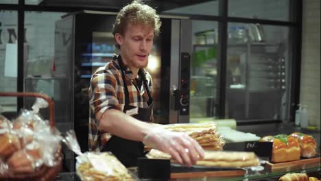 Positive-shop-clerk-in-black-apron-putting-baked-sticks-on-the-showcase-standing-in-the-beautiful-store-with-bakery-products.-Slow-motion