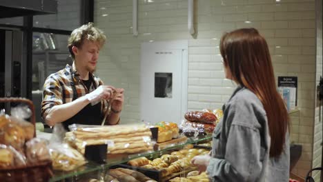Bread-seller-packing-fresh-baguette-for-young-woman-customer-in-the-beautiful-store-with-bakery-products