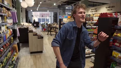 Excited-young-man-in-plaid-shirt-in-supermarket-dancing,-funky-moves-close-to-shopping-trolley.-Positive-dances-in-an-empty-food-store-row.-Positive-emotions.-Slow-motion