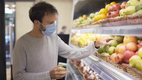 Man-in-mask-takes-a-fresh-apple-from-the-food-shelf.-Shopper-choosing-apple-at-grocery-store