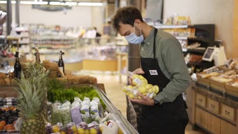 Male-assistant-in-supermarket-food-store-worker-in-medical-mask-and-apron-arranges-packed-products-on-shelves