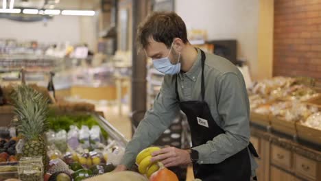 Male-assistant-in-supermarket-food-store-worker-in-medical-mask-and-apron-arranges-bananas-on-shelves