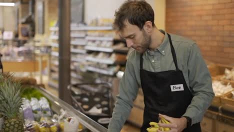 Happy-grocery-store-worker-in-apron-caucasian-bearded-guy-arranging-bananas-at-modern-organic-supermarket-grocery-store