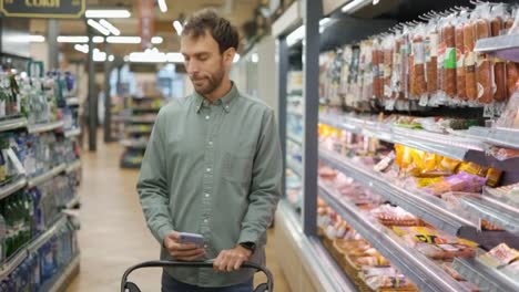 Happy-man-in-shirt-walking-with-a-cart-through-the-supermarket-choosing-groceries.-checking-the-shopping-list-on-the-cell-phone