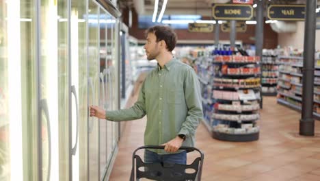 Man-chooses-products-in-the-supermarket.-Opening-refrigerator-and-taking-frozen-vegetables.-Putting-pack-of-frozen-green-beans