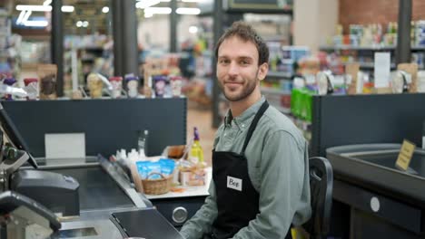 Schöner-Mann-Im-Supermarkt,-Der-An-Der-Kasse-Auf-Den-Nächsten-Kunden-Wartet.-Lächelnde-Kassiererin-In-Schwarzer-Schürze