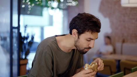 Unshaven-c-man-eats-meat-burger-with-pleasure,-bites-burger-with-his-mouth