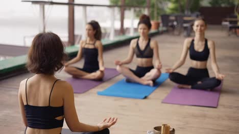 Group-of-girls-sitting-on-the-outdoors-wooden-floor-in-lotus-pose,-yoga-led-by-trainer