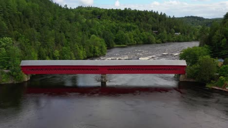 Aerial-dolly-shot-toward-a-bright-red-covered-timber-bridge-over-a-Canadian-river-with-rapids-downstream