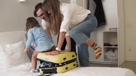 Young-caucasian-couple-tries-to-zip-suitcase-full-of-clothes.-Parents-closing-suitcase-and-ask-their-little-daughter-to-sit-on