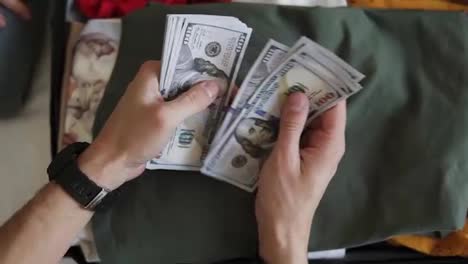 Unrecognizable-man-hands-with-pile-of-money,-US-dollars-notes---counting-under-the-suitcase-with-clothes.-Counting-money-for