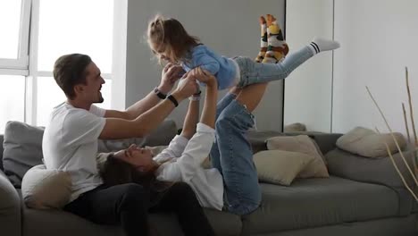 Happy-family-mom-and-dad-having-fun-with-little-girl-on-cozy-home-grey-sofa.-Little-daughter-imitates-flying-being-on-her