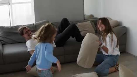 Little-cute-girl-having-funny-pillow-fight-with-dad-on-couch-in-living-room-and-mom-is-sitting-on-the-floor-playing-with-girl
