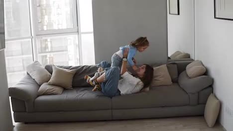 Happy-family-mother-and-kid-daughter-having-fun-together-lying-on-sofa.-Young-woman-tickles-the-girl,-raises-in-outstretched