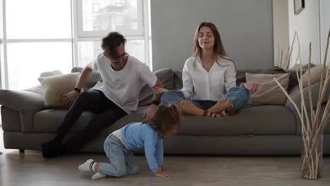 Mindful-caucasian-mother-meditating-sitting-on-sofa-while-active-energetic-child-daughter-jumping-playing-around-and-father-sits