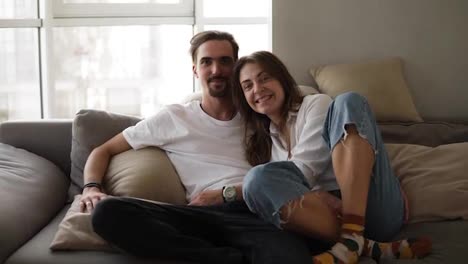 Portrait-shot-of-the-young-caucasian-smiled-bearded-boyfriend-and-girlfriend-sitting-on-the-grey-sofa-and-posing-to-the-camera