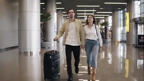 Front-view-of-smiling-couple-walking-together-in-airport-going-on-vacation-or-trip.-Travel-together.-Attractive-caucasian-young