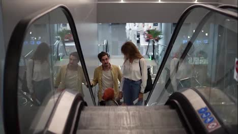 Young-attractive-caucasian-family-with-suitcase-going-up-by-the-escalator-with-their-pretty-daughter-hold-by-mother's-hand-at-the